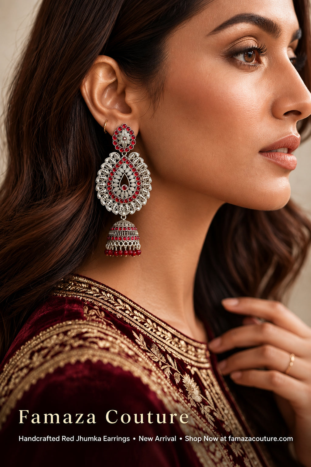 A close-up side profile of a woman wearing a large, ornate Famaza Couture handcrafted red Jhumka earring. The earring features silver-toned metalwork, clear stones, and vibrant red accents, paired with a deep maroon embroidered traditional outfit.